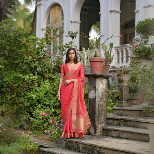A woman models a stunning Coral Red Soft Silk Saree. The sheer saree features subtle metallic motifs and a wide, contrasting metallic gold/bronze woven border and pallu. She wears a matching red short-sleeve blouse and delicate gold jewelry, posing gracefully on an ornate stone staircase.