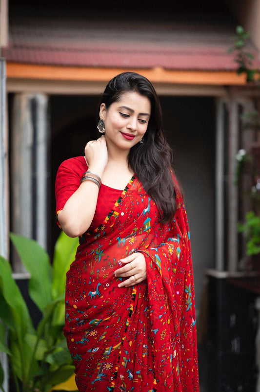 Woman in a red Warli print saree with pom-pom border standing on a tiled patio with lush greenery and a traditional house in the background