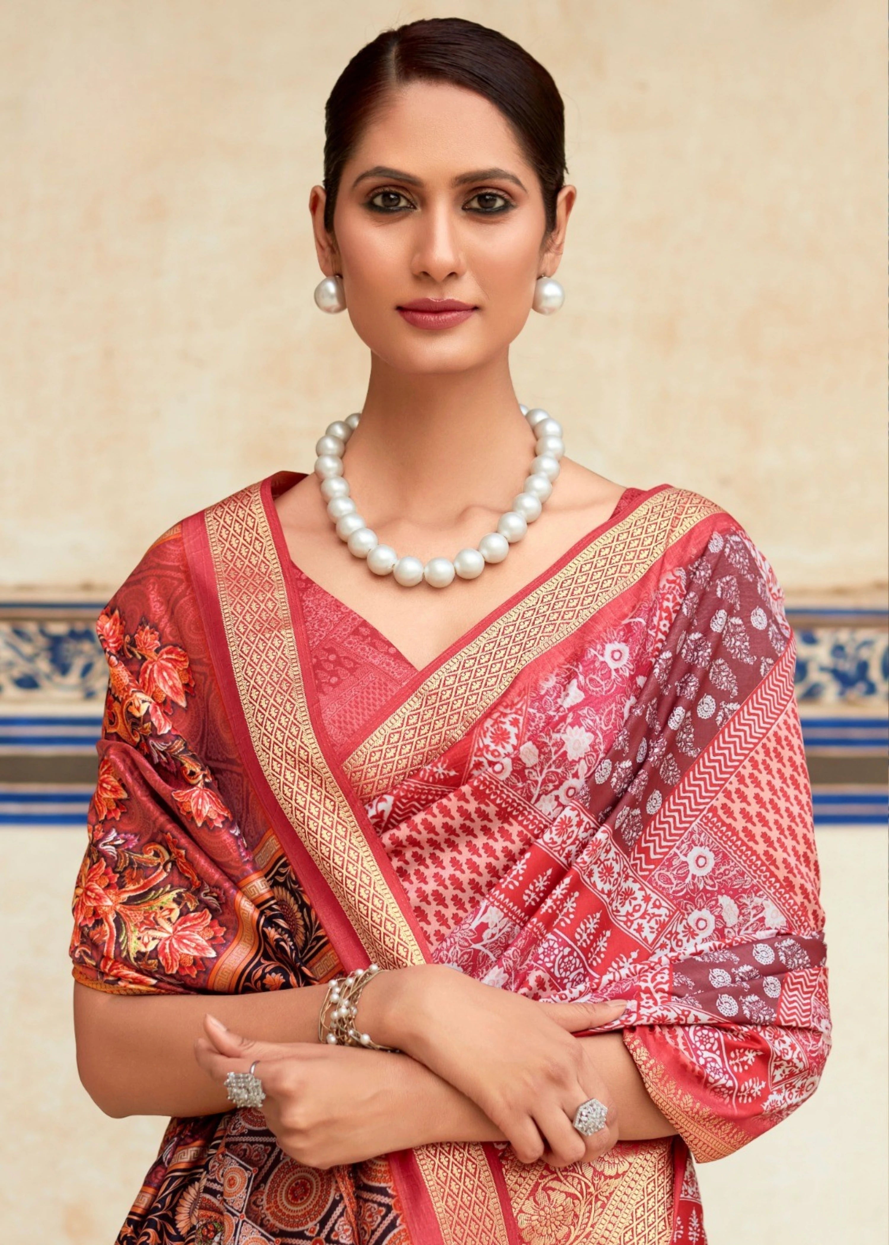 Woman wearing a red saree with floral patterns and a pearl necklace against a decorative wall.