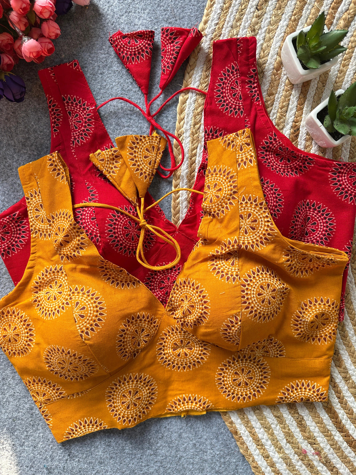 Red and yellow embroidered tops on a textured surface with flowers and plants in the background.