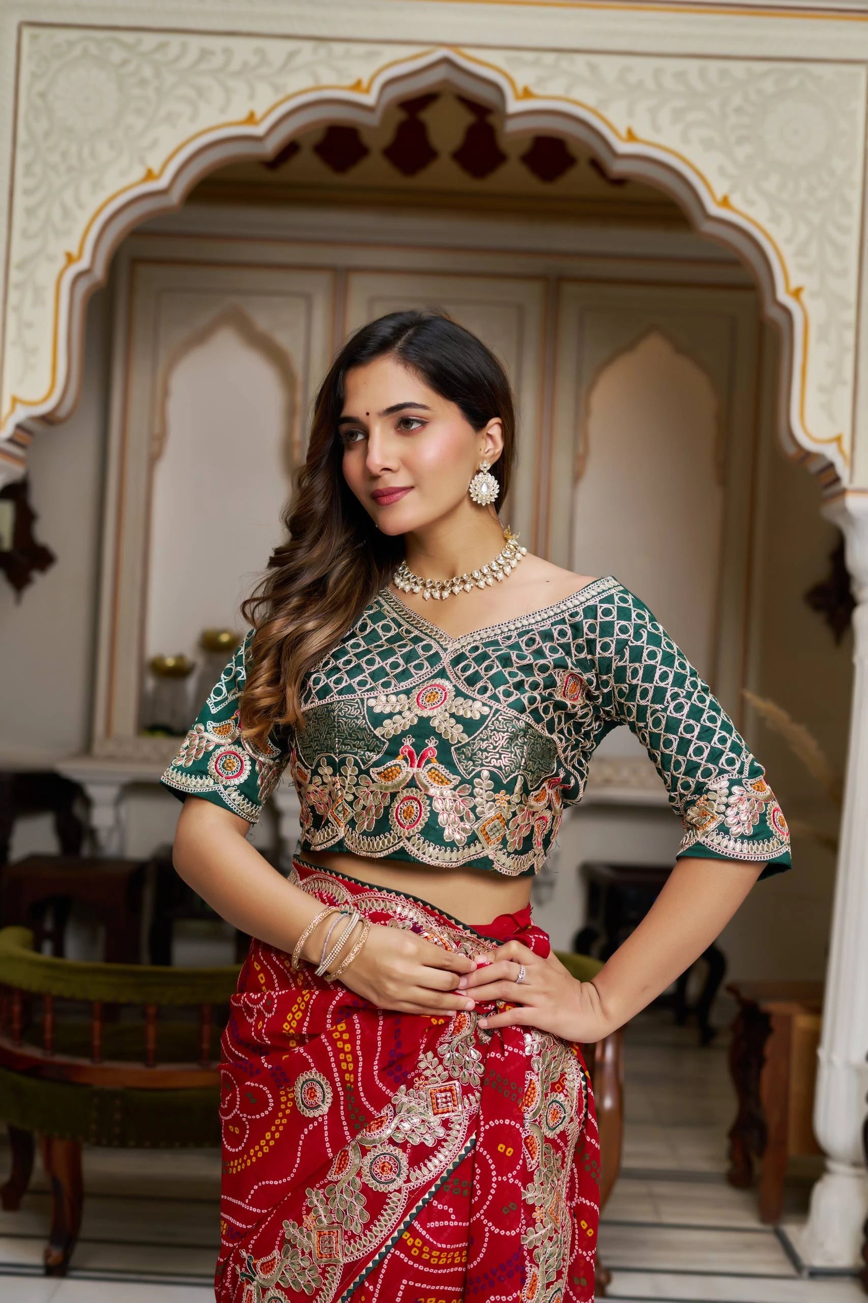 Woman wearing a traditional outfit with a green blouse and red saree in an ornate room.