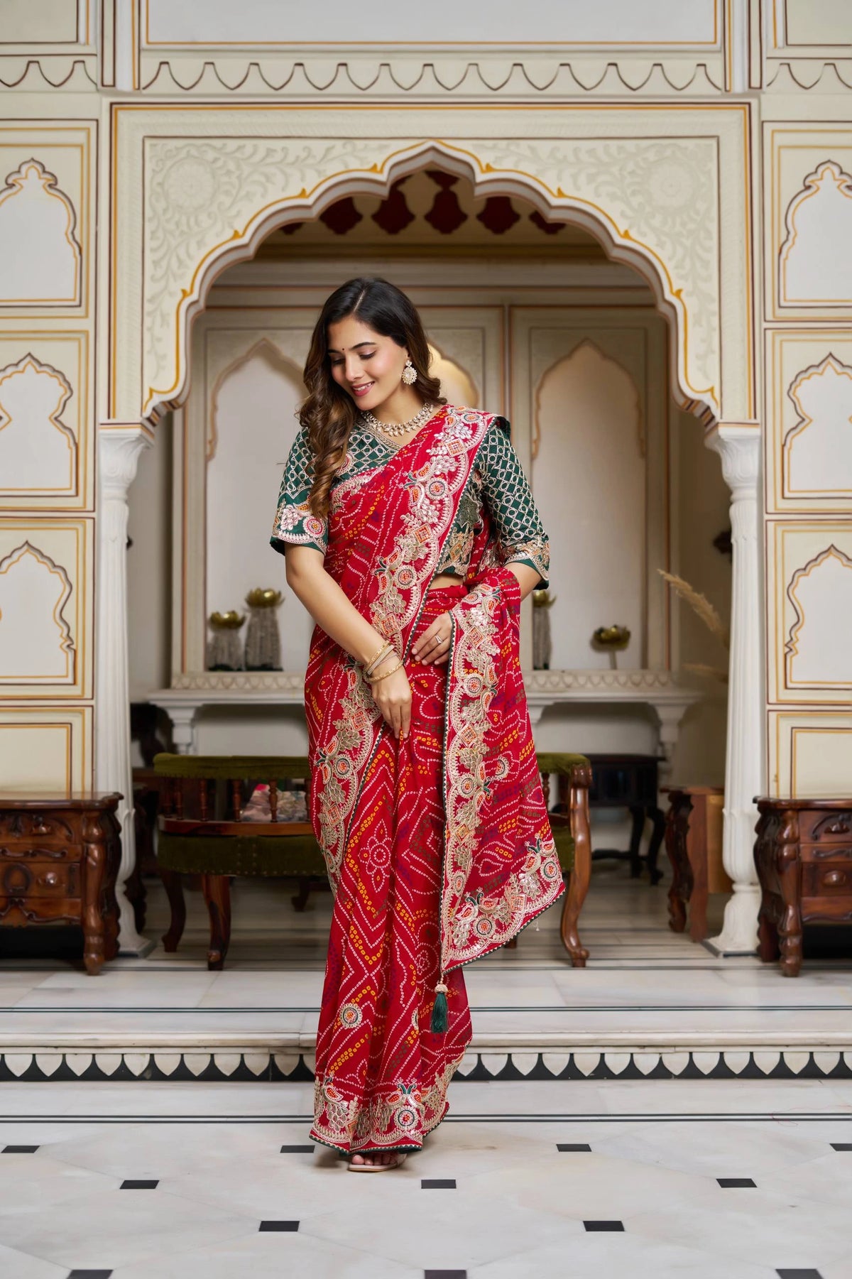 Woman in a red saree with gold patterns standing in an ornate room.