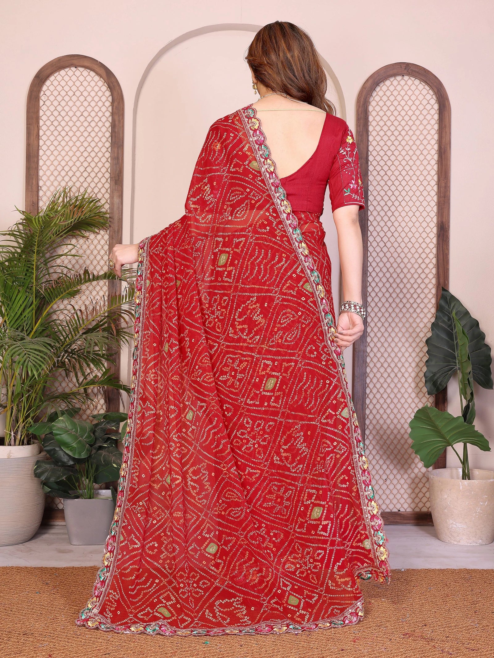 Woman wearing a red saree with a pattern, standing indoors with plants in the background.