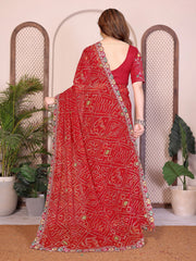 Woman wearing a red saree with a pattern, standing indoors with plants in the background.