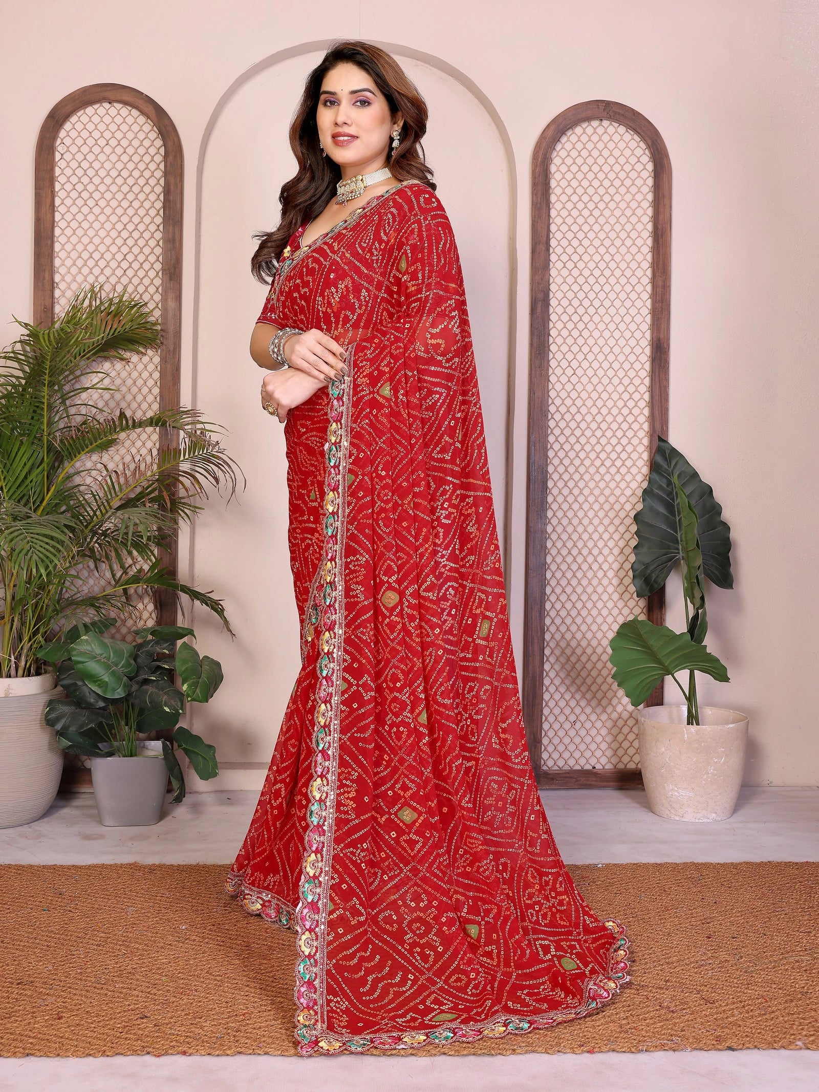 Woman wearing a red saree with intricate patterns in a decorative setting with plants.