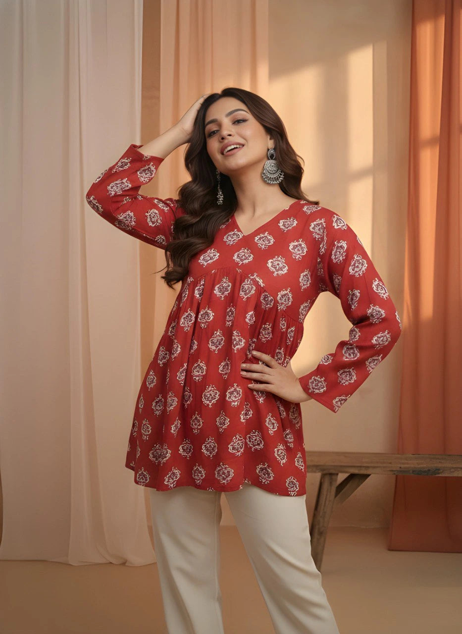 Woman wearing a red patterned top and white pants in a warm-toned room.