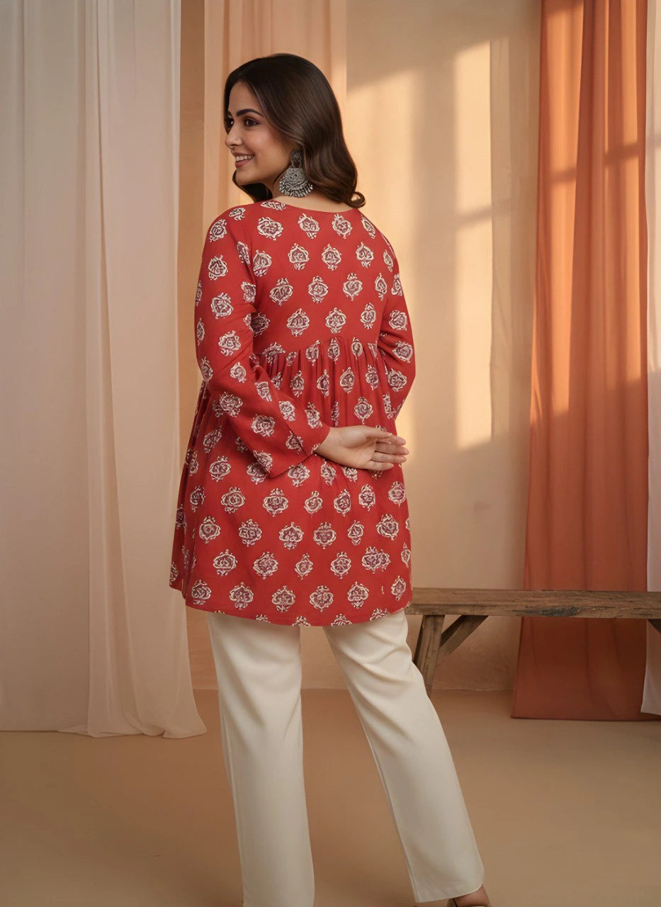 Woman wearing a red patterned top and white pants standing in a room with orange curtains.
