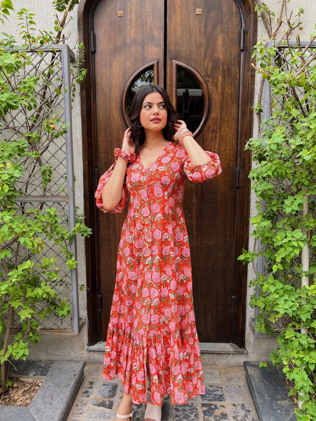 Woman in a red floral dress standing in front of a wooden door with greenery around.