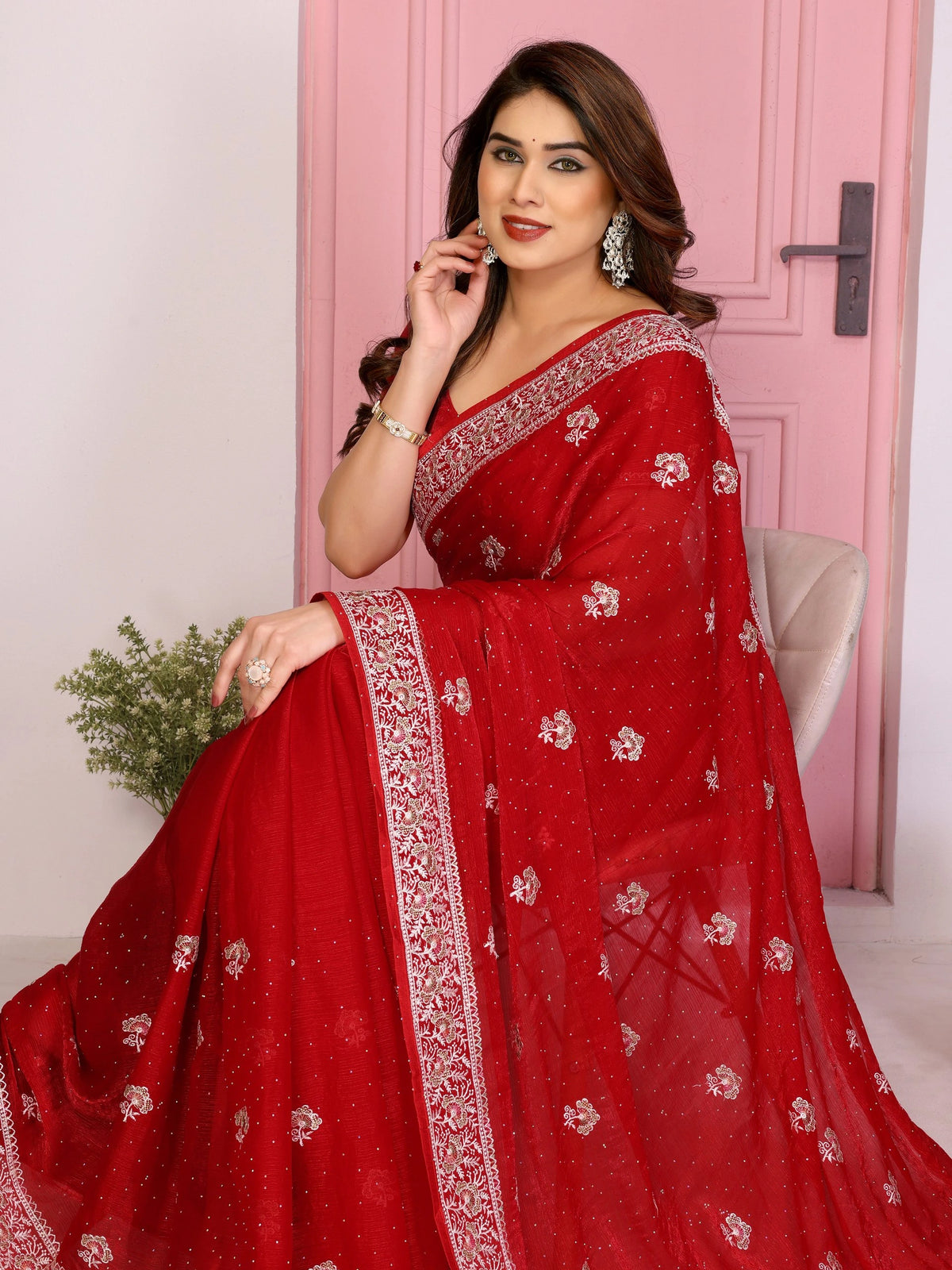A medium shot of a woman sitting gracefully on a chair, draped in a red saree with detailed silver embroidery. She is smiling softly while looking toward the side, highlighting the delicate texture and shimmer of the fabric.