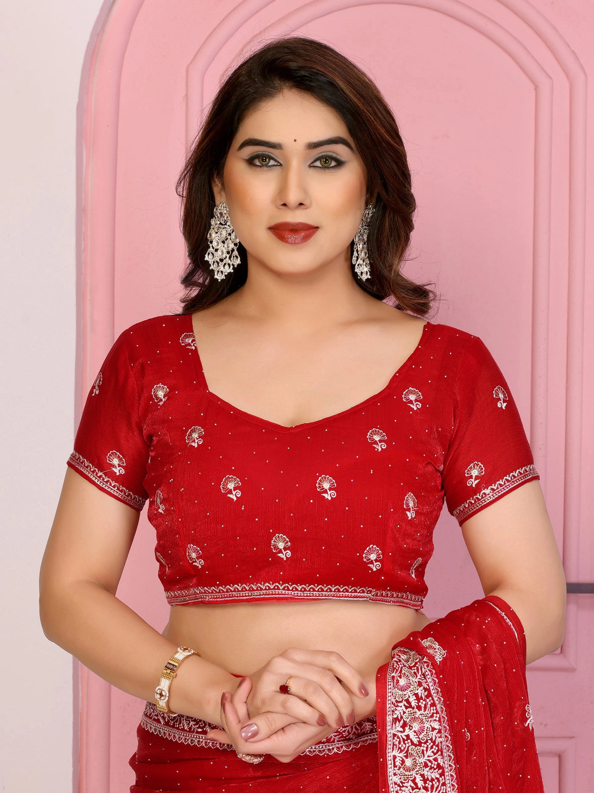 A close-up portrait of a woman wearing a red blouse with intricate silver floral embroidery and a matching red saree. She is adorned with large, ornate silver drop earrings and a gold bracelet, with her hands clasped in front