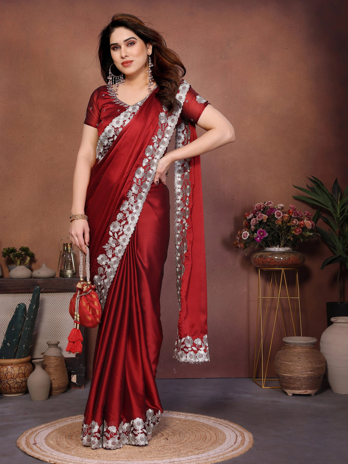 Woman in a red saree with white border posing in a room with decorative plants and vases.