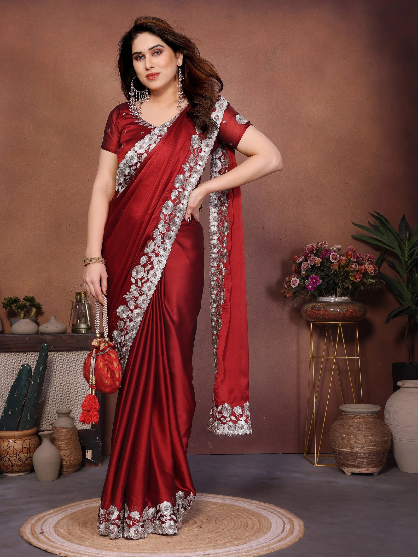 Woman in a red saree with white border posing in a room with decorative plants and vases.