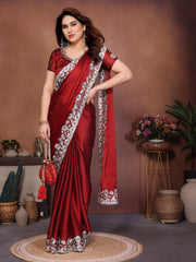 Woman in a red saree with white border posing in a room with decorative plants and vases.