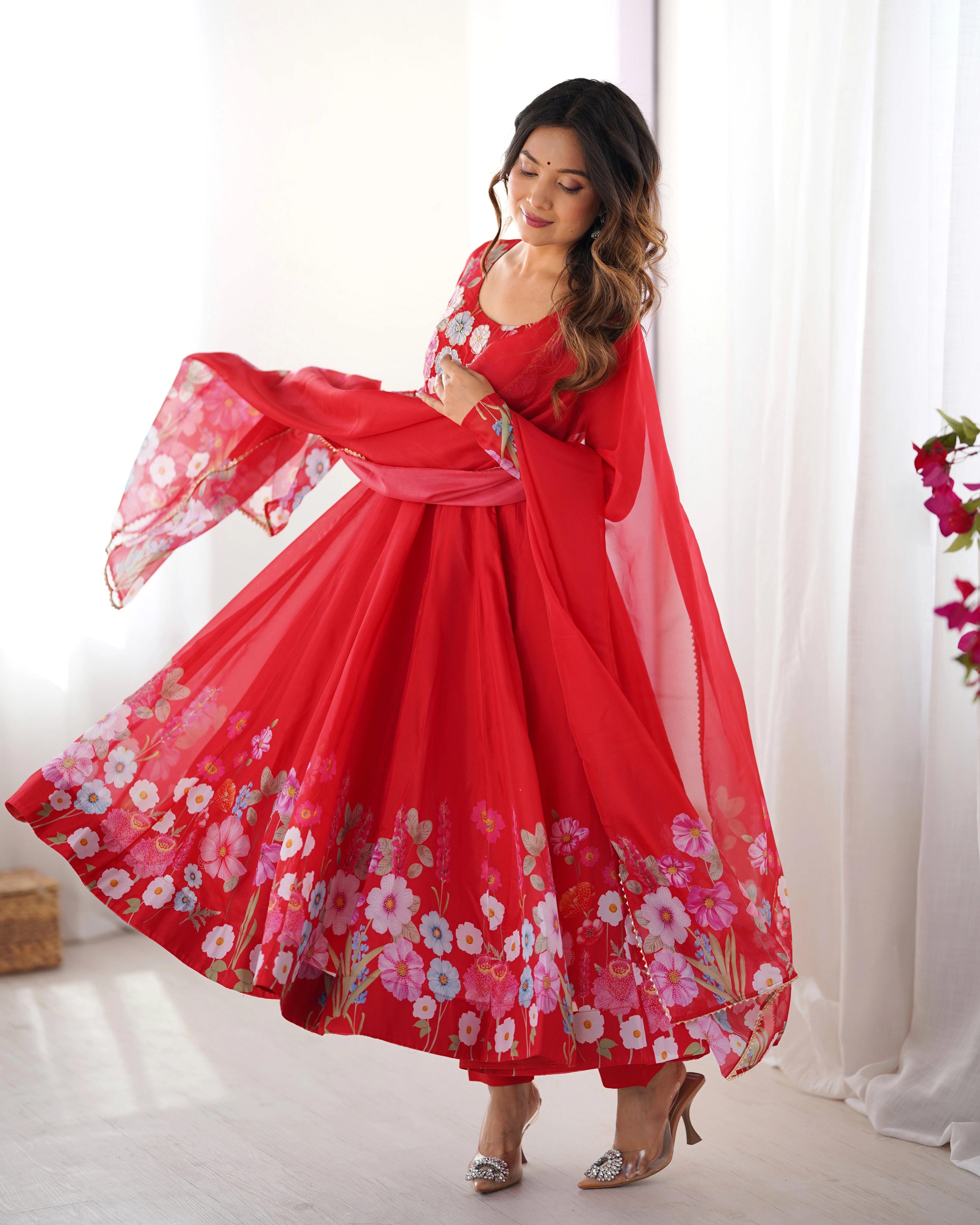 Woman in a red floral dress posing in a bright room with white curtains.