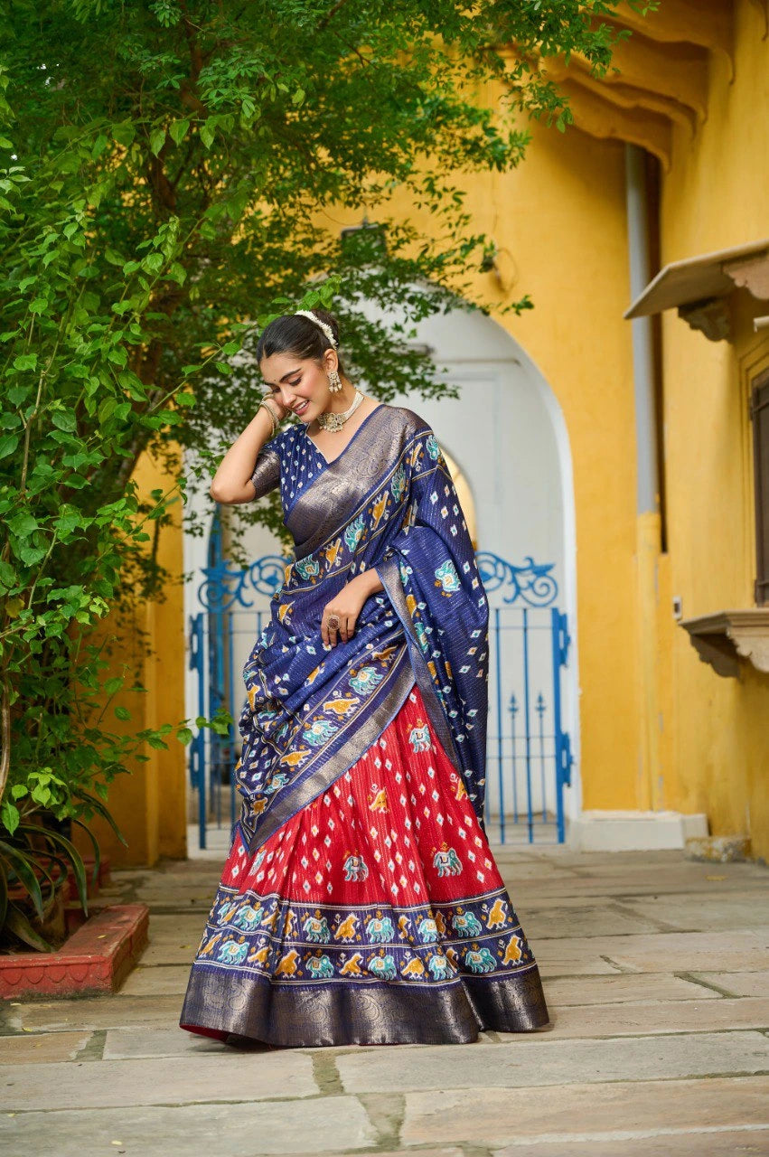 Woman in a traditional blue and red saree standing in front of a yellow wall with an archway.
