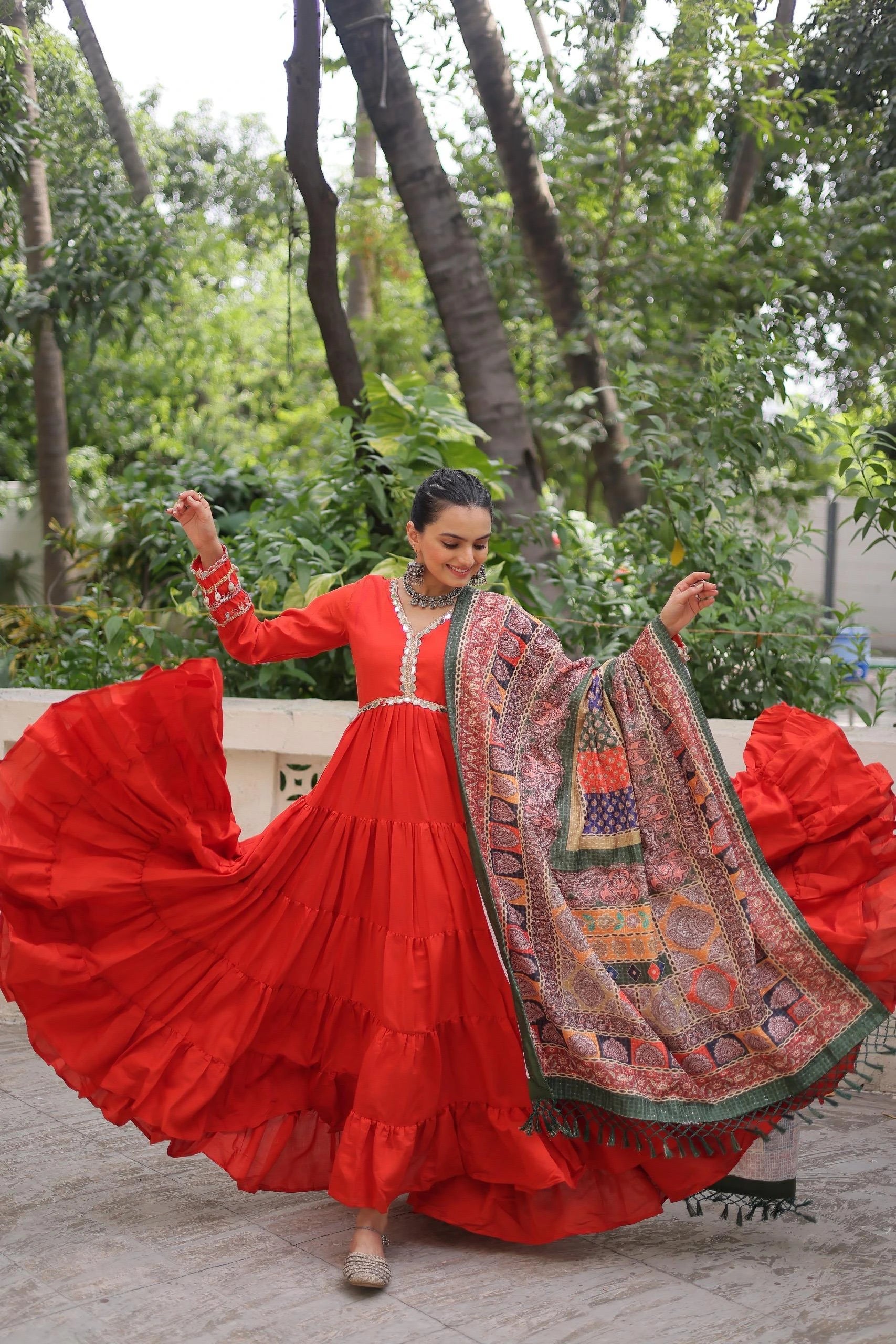 Woman in a red traditional outfit with a patterned shawl, standing outdoors with greenery in the background.