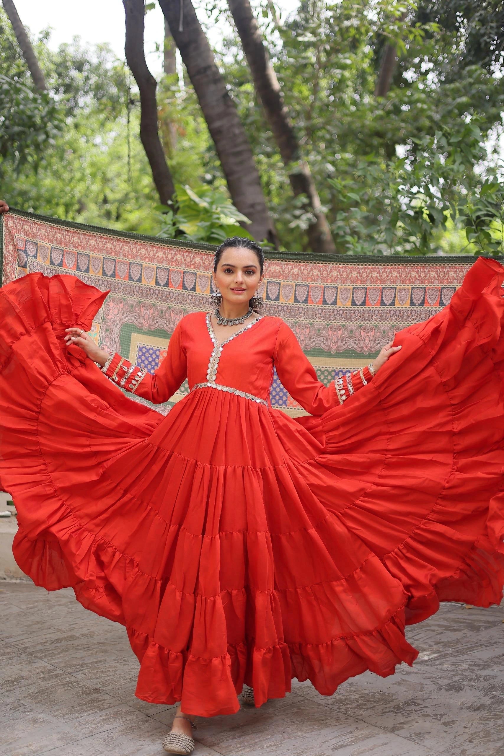 Woman in a red traditional outfit with a flowing skirt, standing outdoors with greenery in the background.