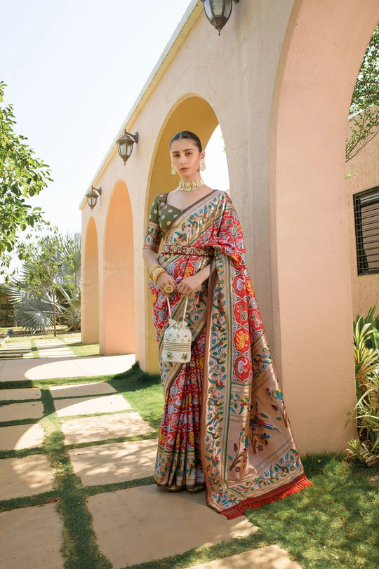 Woman in a colorful saree standing outdoors near a building with greenery.
