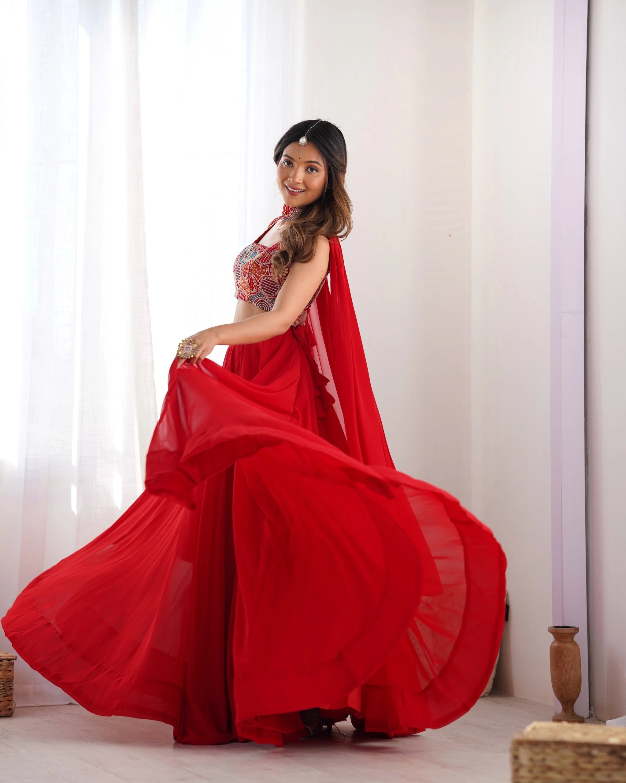Woman in a red saree standing in a well-lit room with white curtains.