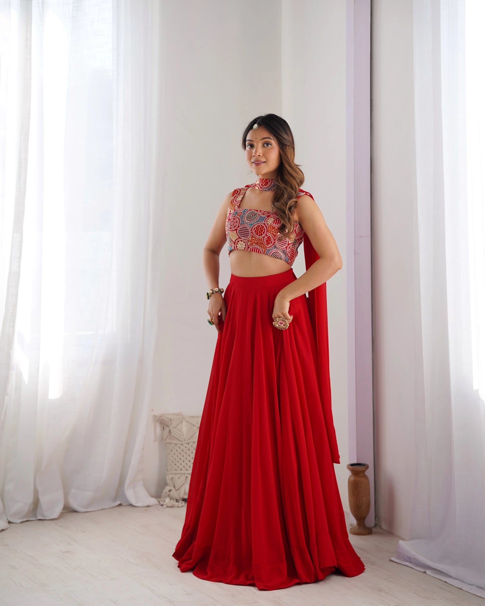 Woman wearing a red and patterned top with a long skirt against a white curtain background