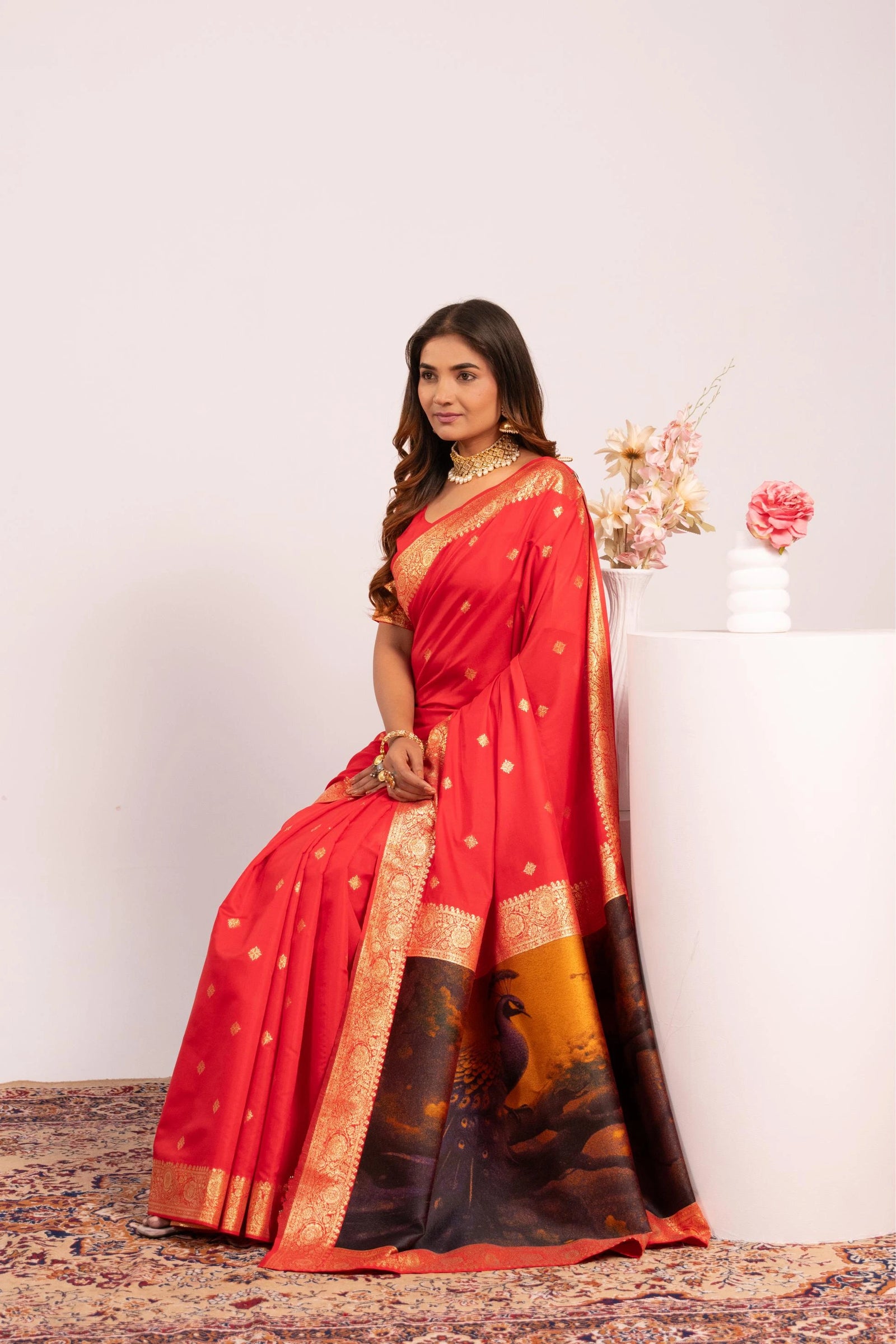 Woman wearing a red saree with gold border sitting on a white pedestal.