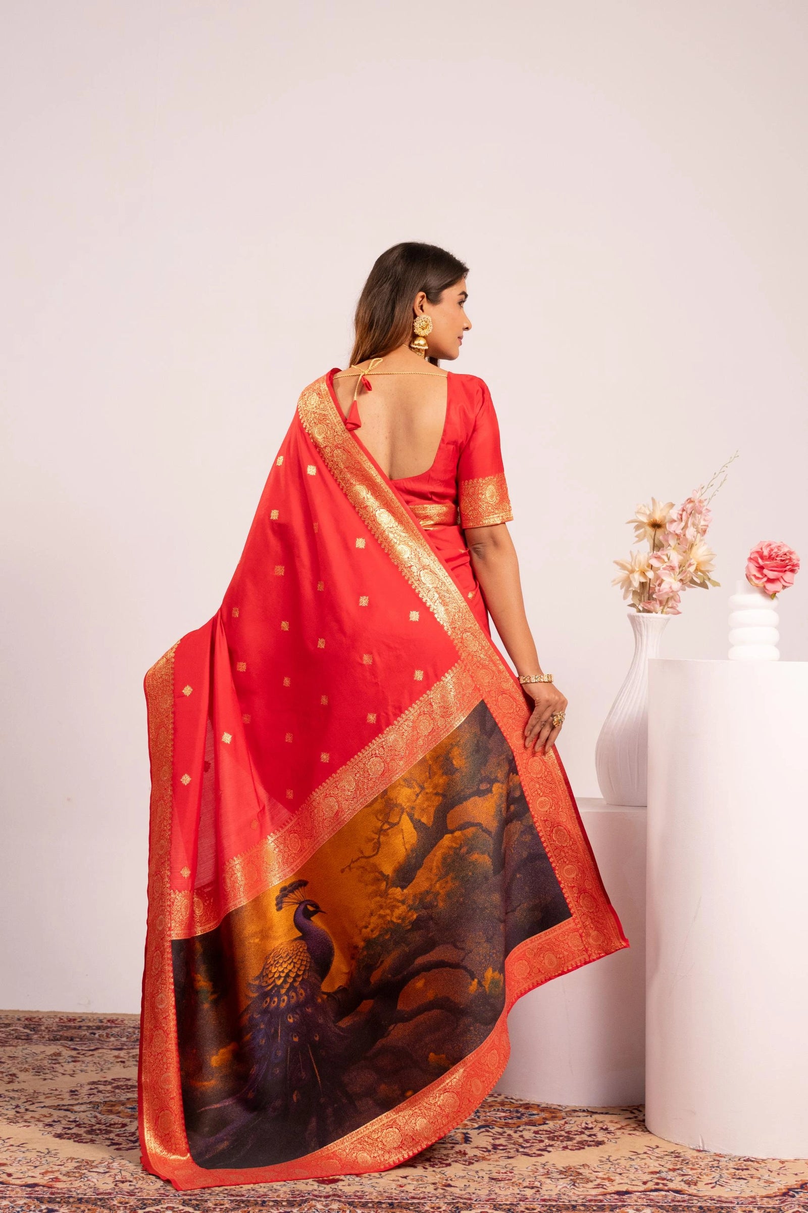 Woman wearing a red saree with peacock design, standing against a white wall.