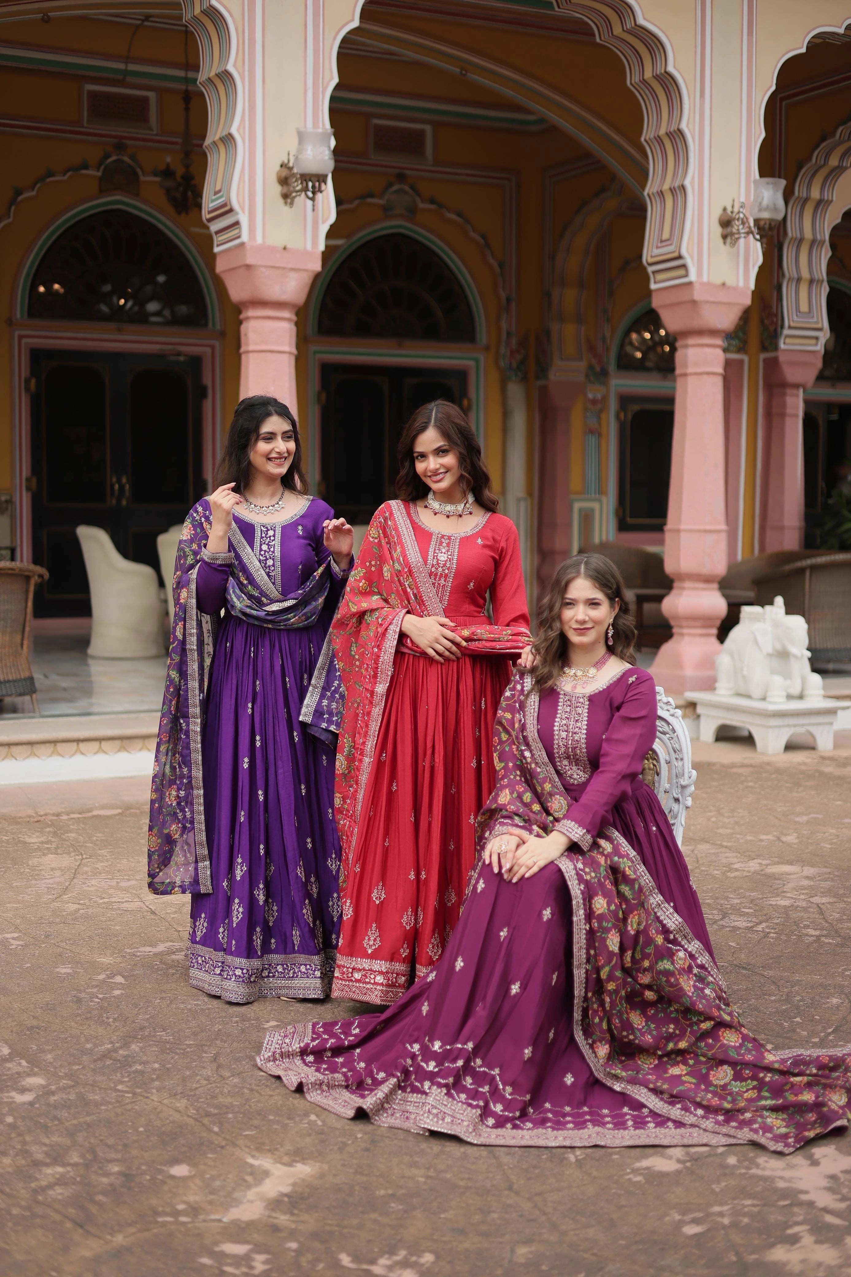Three women in traditional purple and red dresses posing in front of a pink architectural building.