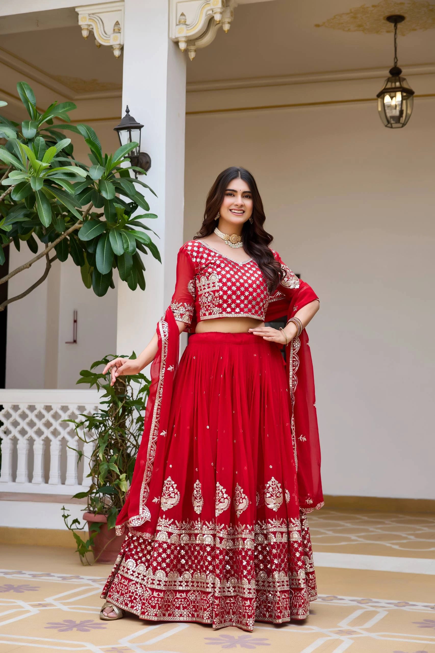Woman in a red traditional outfit standing in an elegant indoor setting with plants and architectural elements.