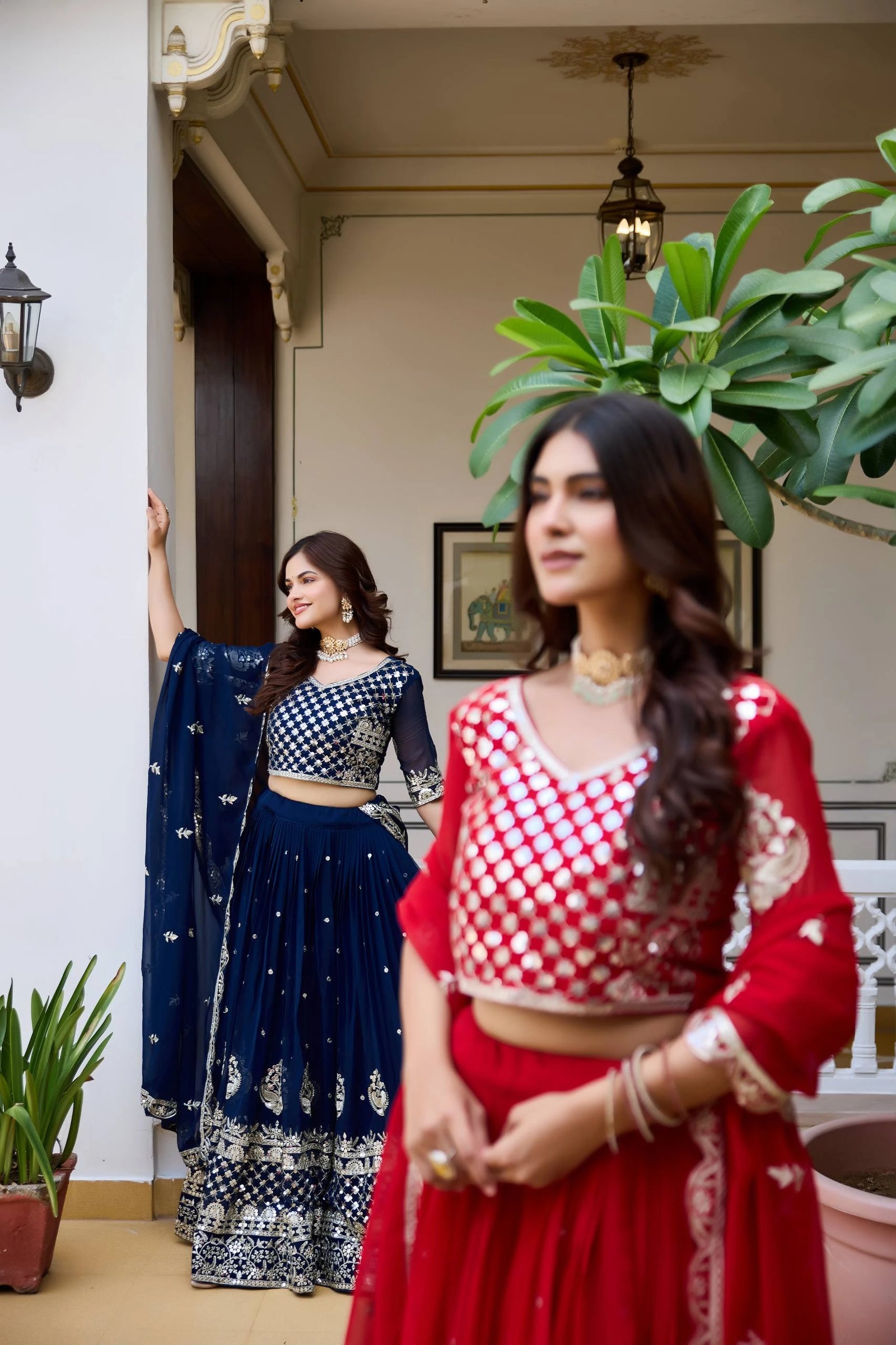 Two women in traditional outfits standing in a room with plants and decor.