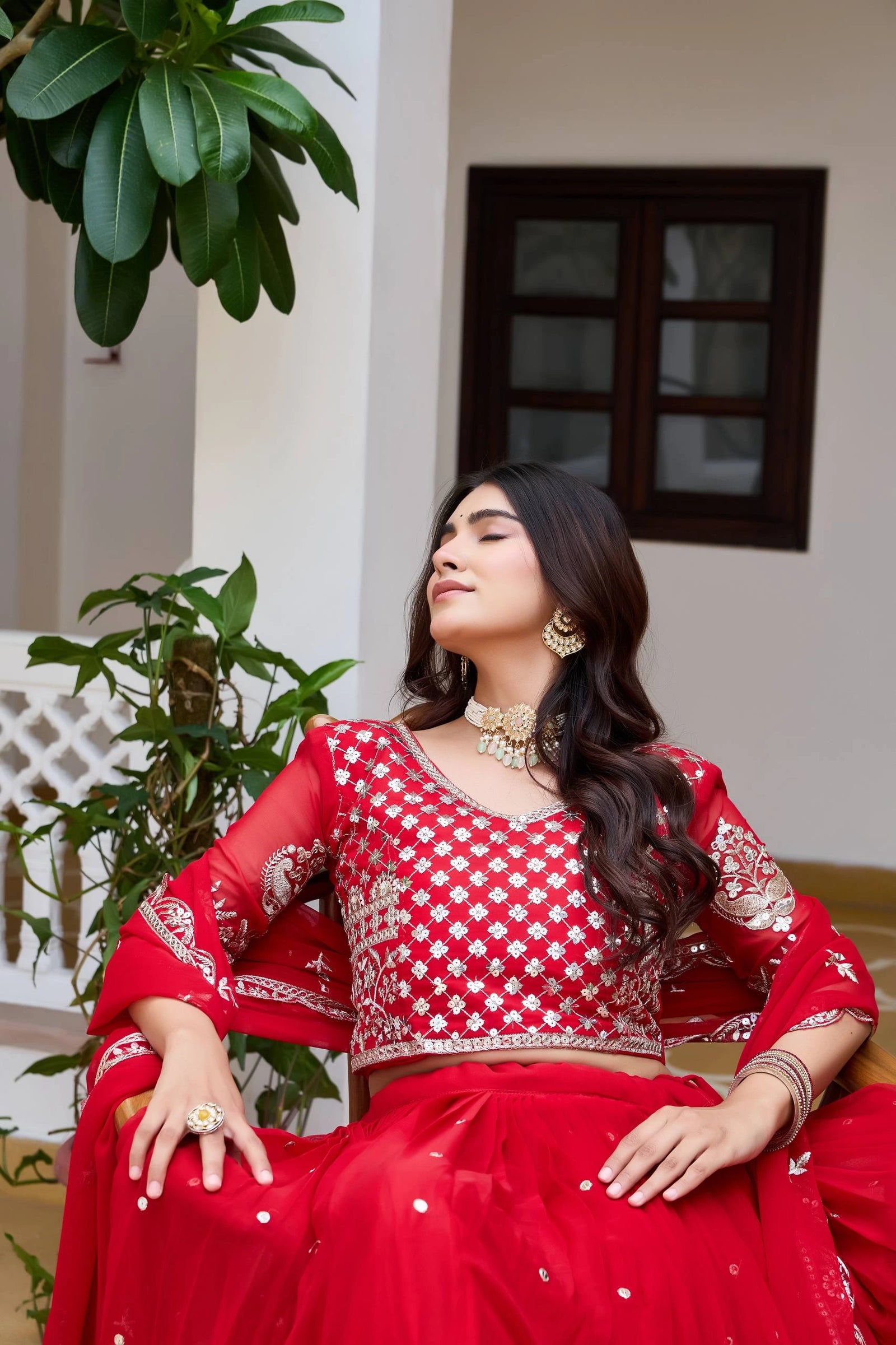 Woman in a red traditional outfit sitting indoors with plants around