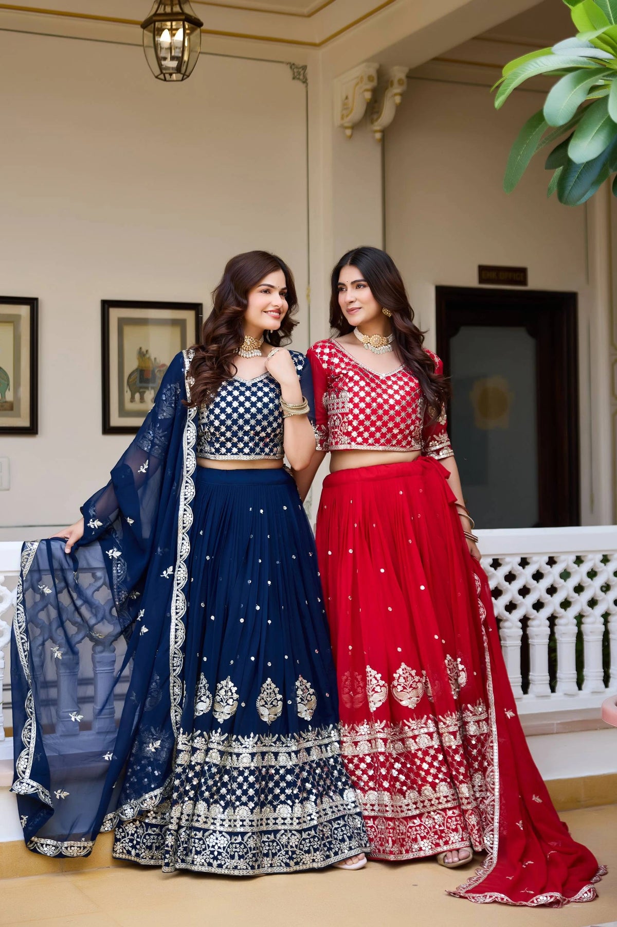 Two women in traditional blue and red lehengas standing indoors.