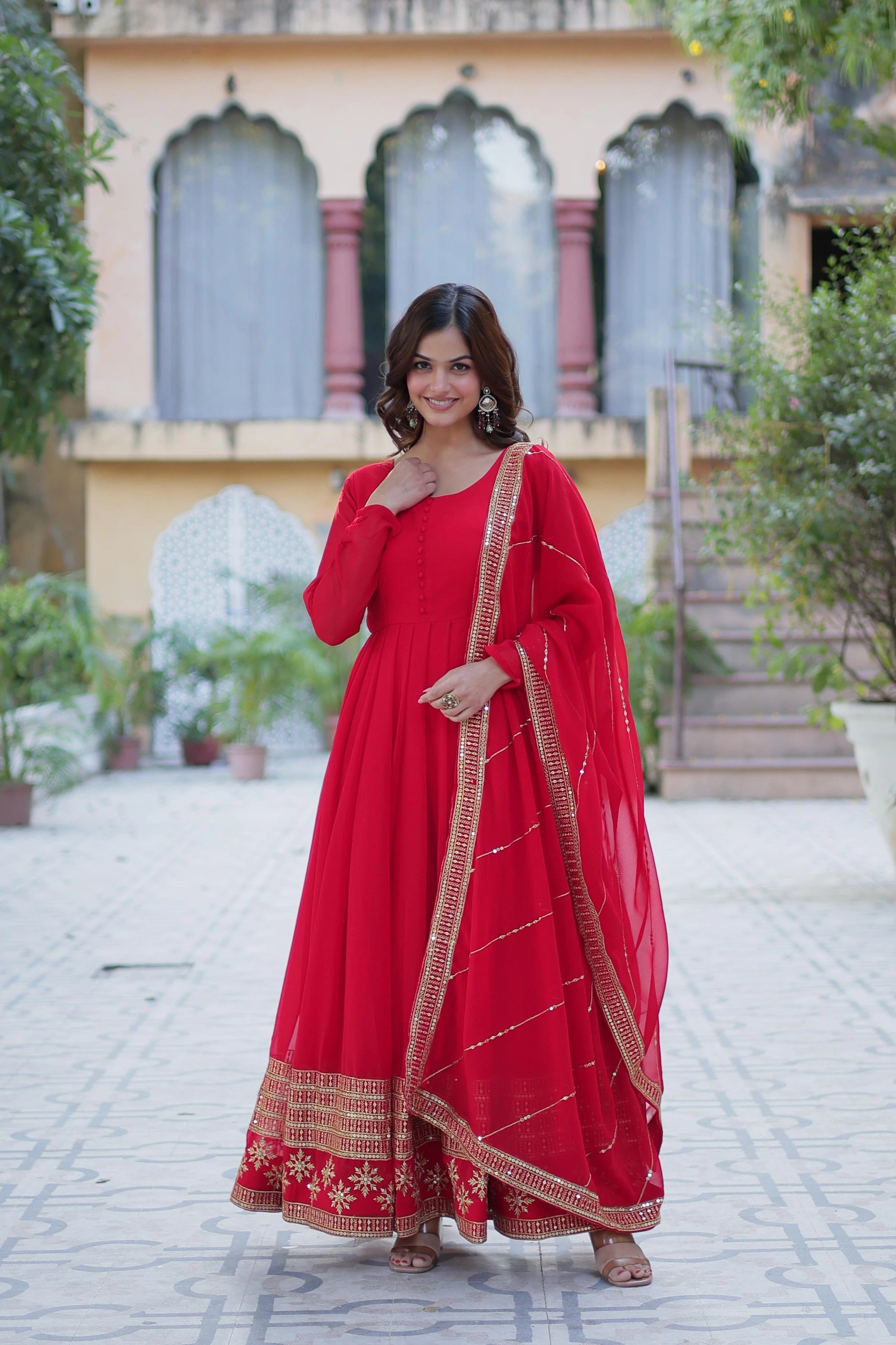 Woman in a red traditional outfit standing outdoors with a building and plants in the background