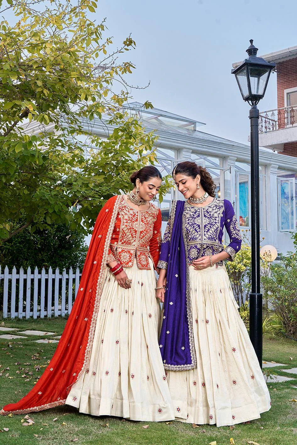 Two women in traditional outfits standing outdoors with a garden and building in the background.