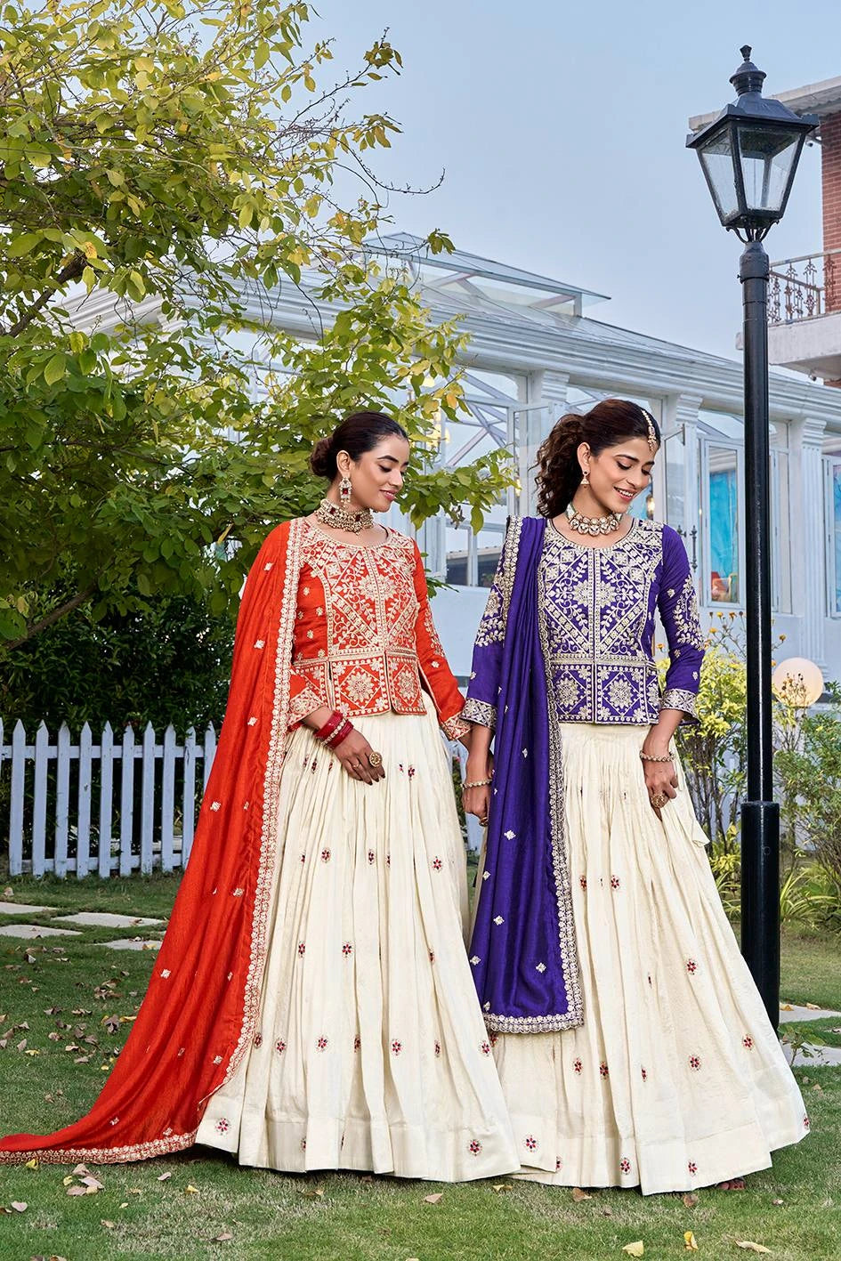 Two women in traditional outfits standing outdoors with a building and trees in the background.