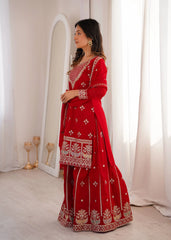 Woman in a red traditional outfit with white patterns standing in a softly lit room.