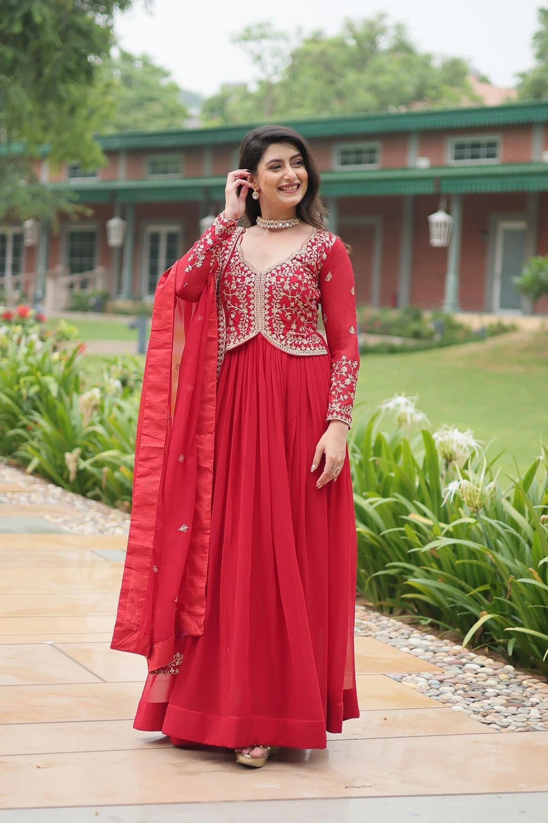Woman in a red traditional outfit standing outdoors with greenery and a building in the background