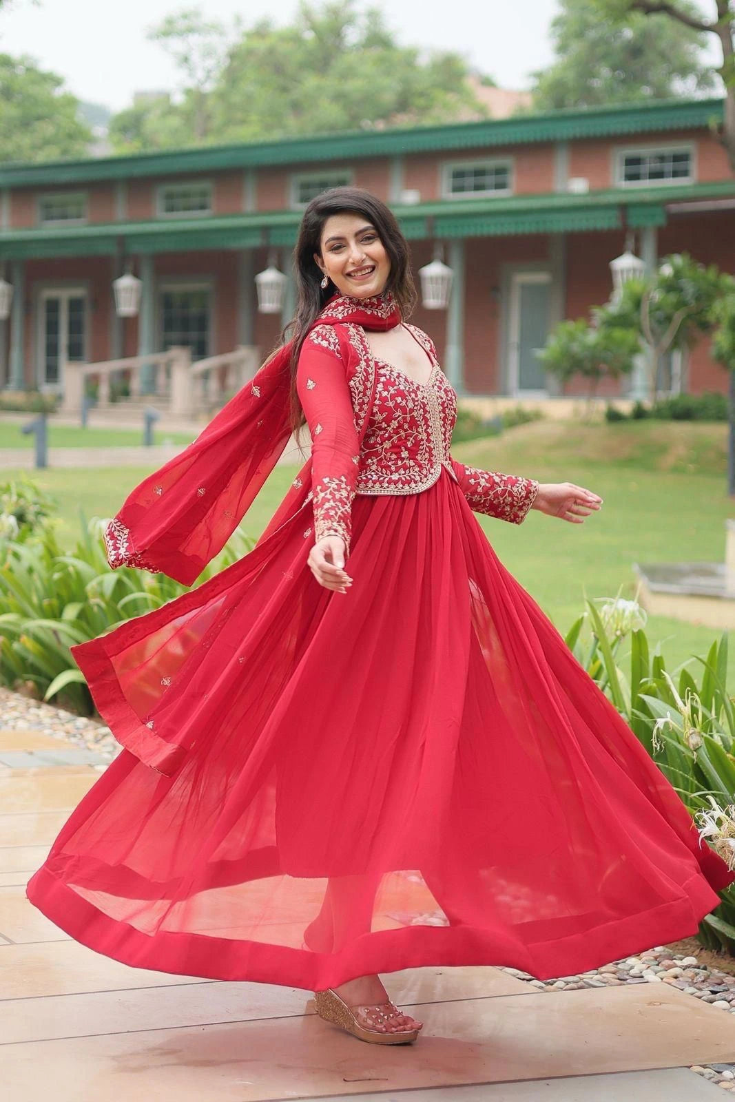 Woman in a red traditional outfit standing outdoors with a building and greenery in the background
