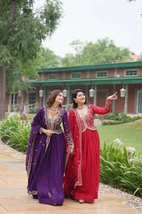 Two women in traditional purple and red dresses standing outdoors with greenery and a building in the background.