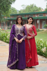 Two women in traditional purple and red dresses standing outdoors with greenery and a building in the background.