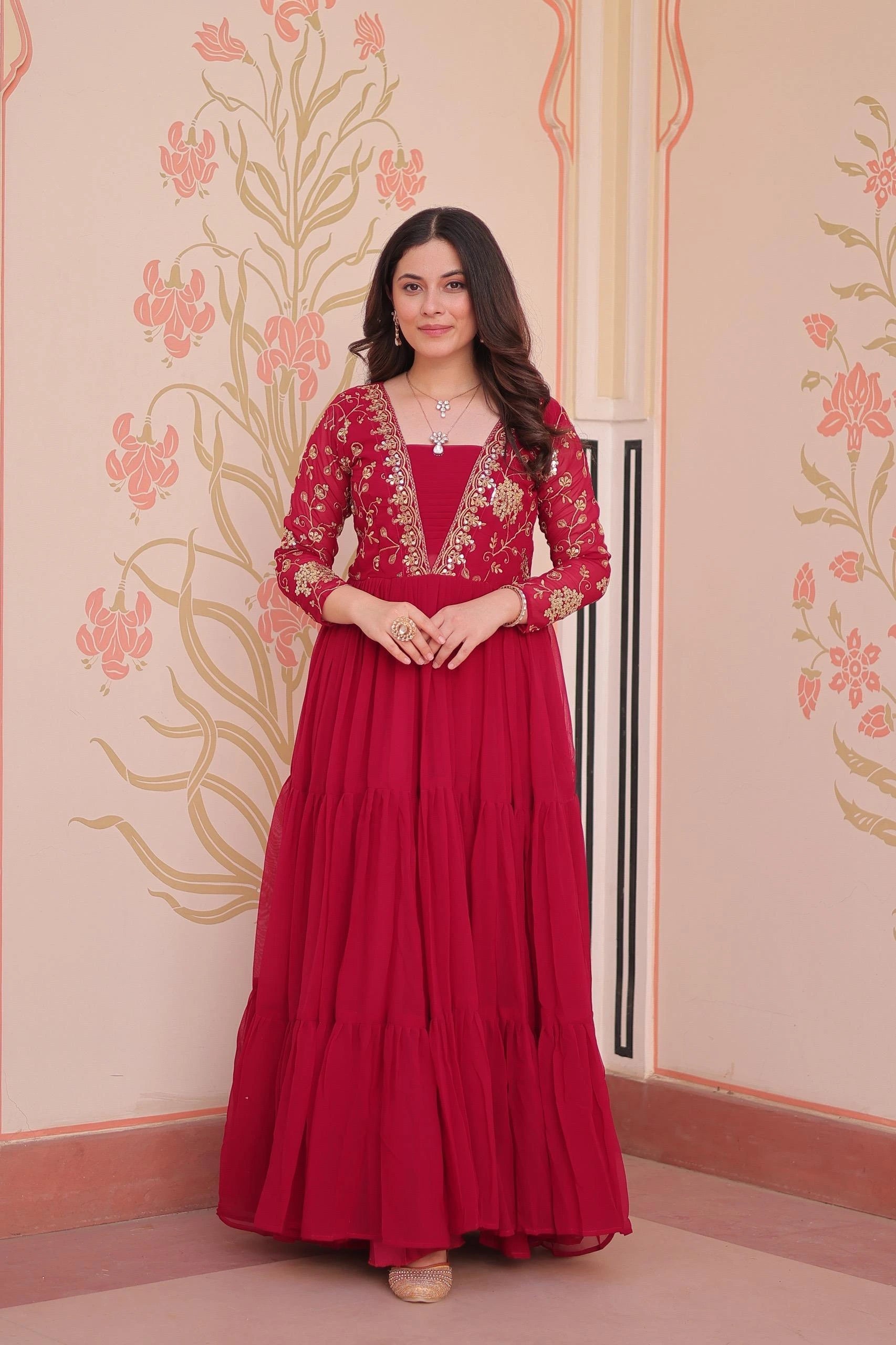 Woman wearing a red traditional dress with intricate designs against a decorative wall.