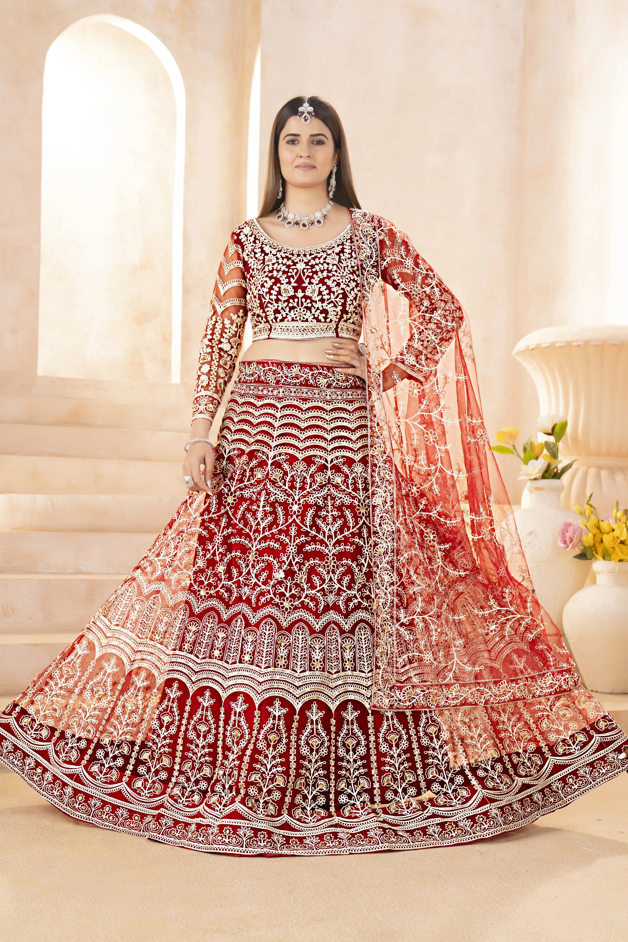 Woman in a red and white embroidered traditional outfit with a matching dupatta, standing indoors.