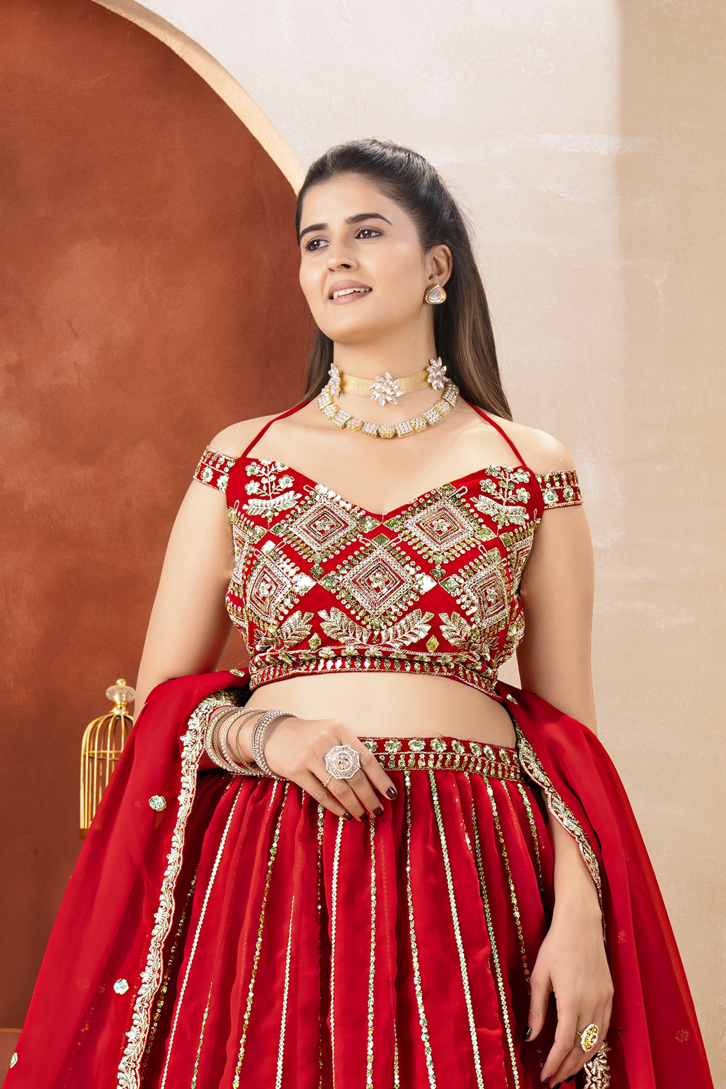 Woman in a red and gold traditional outfit with jewelry against a beige wall.