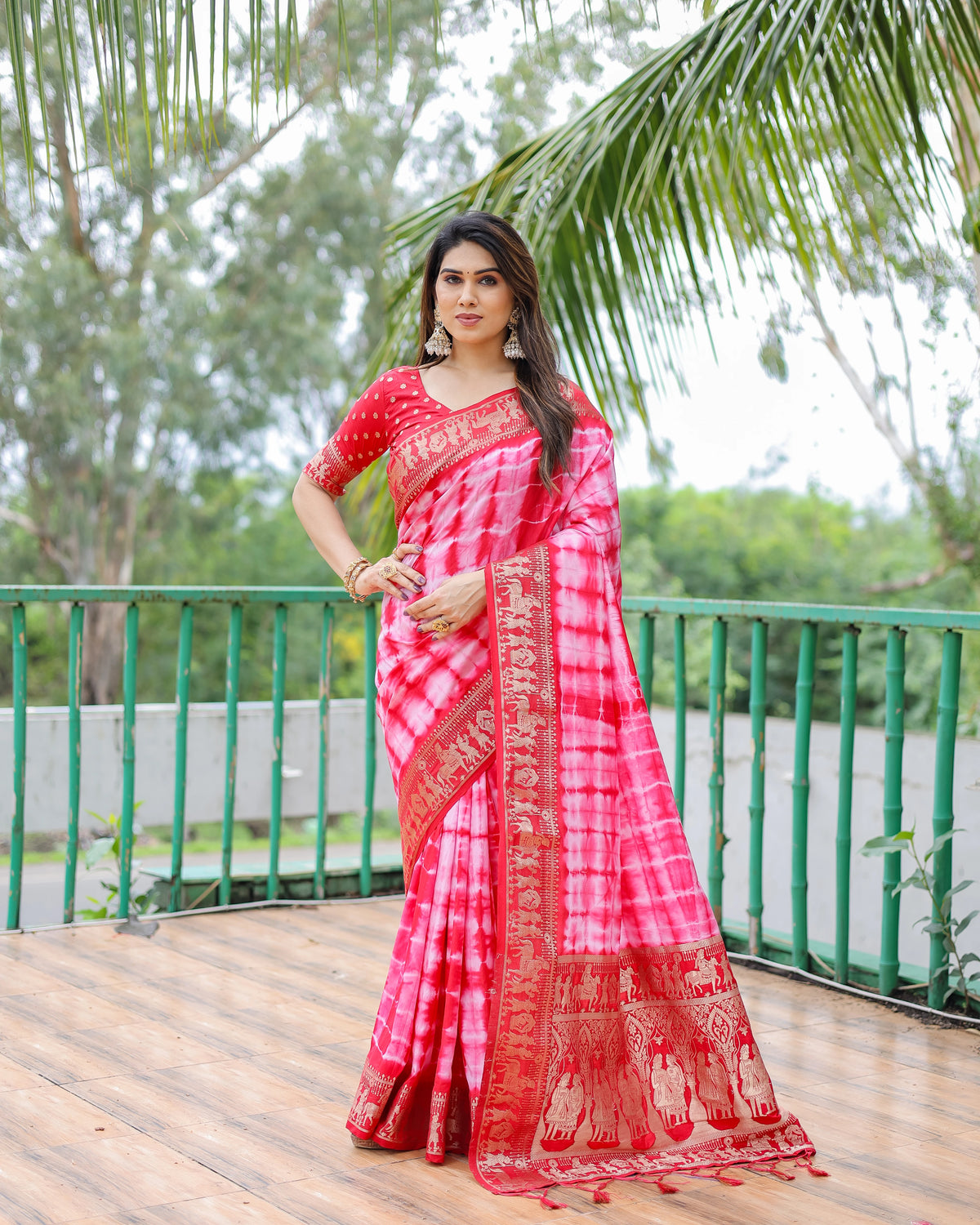 Woman in a red and pink saree standing outdoors with greenery in the background