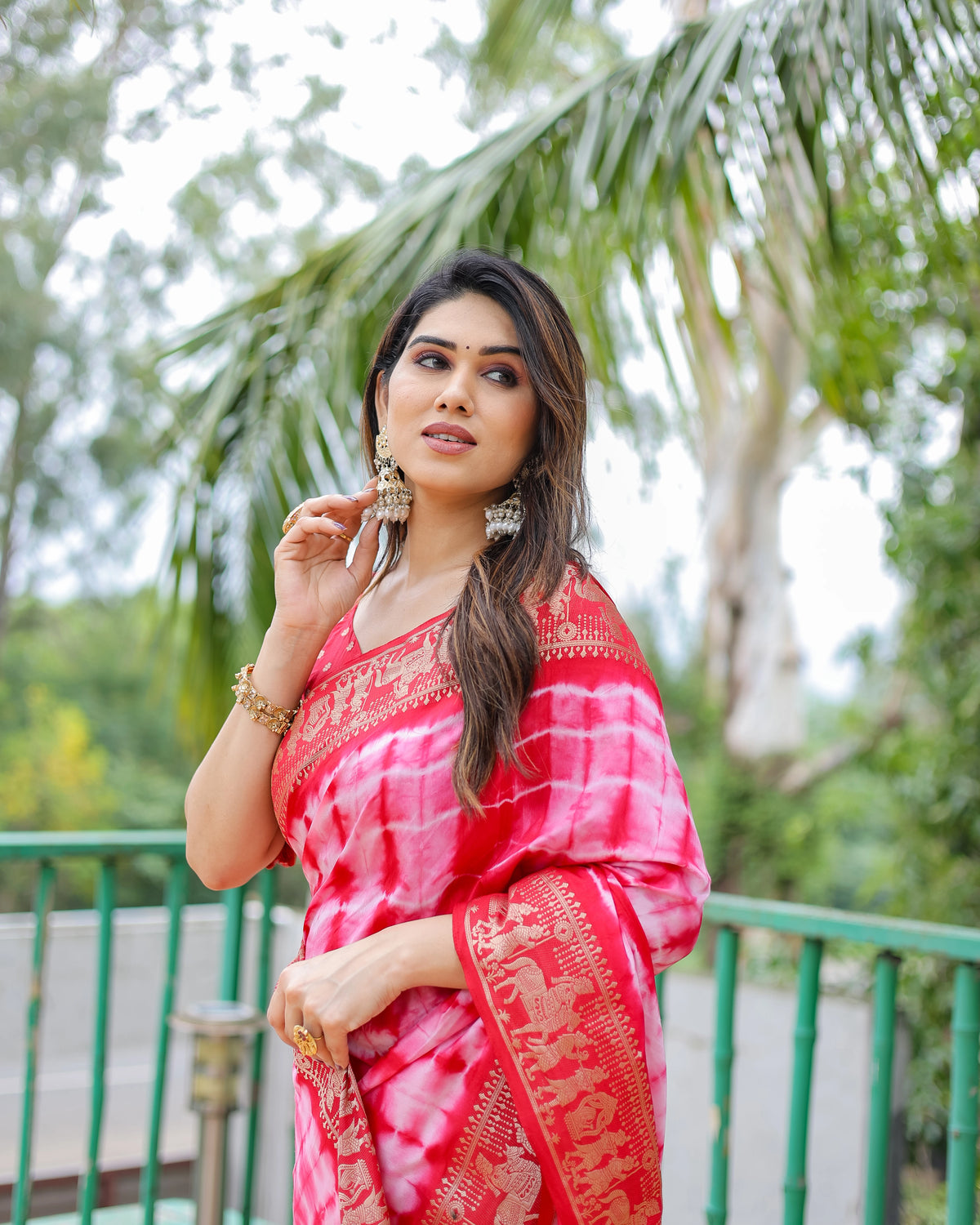Woman in a pink saree with gold patterns standing outdoors with greenery in the background