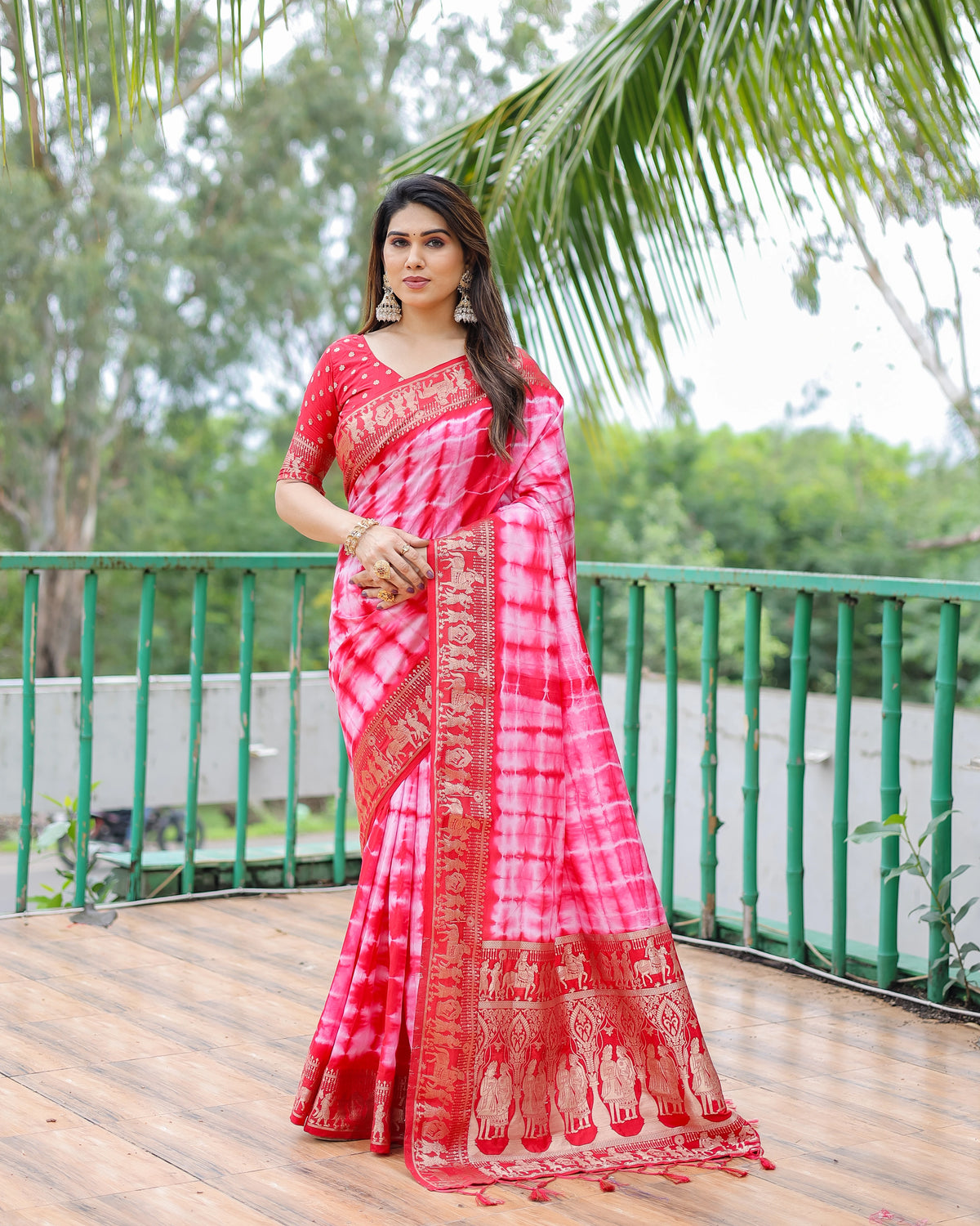 Woman in a red and white patterned saree standing on a wooden deck with greenery in the background