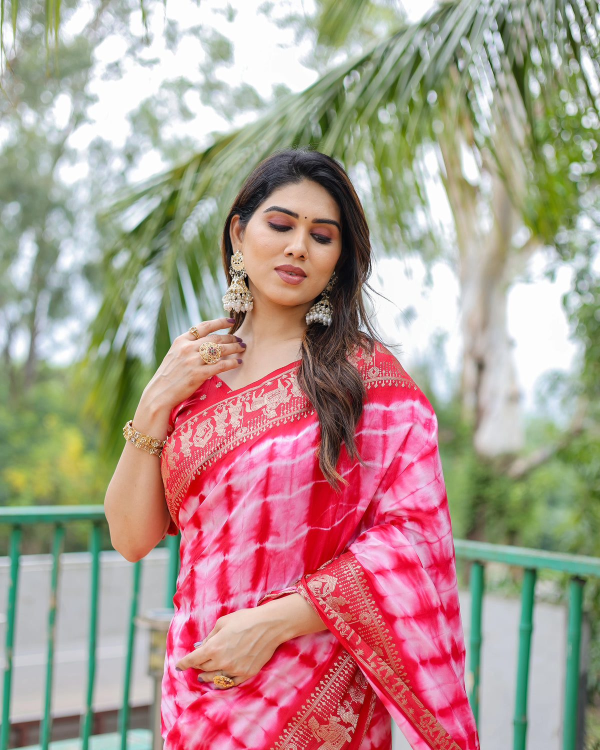 Woman in a red and white saree standing outdoors with greenery in the background