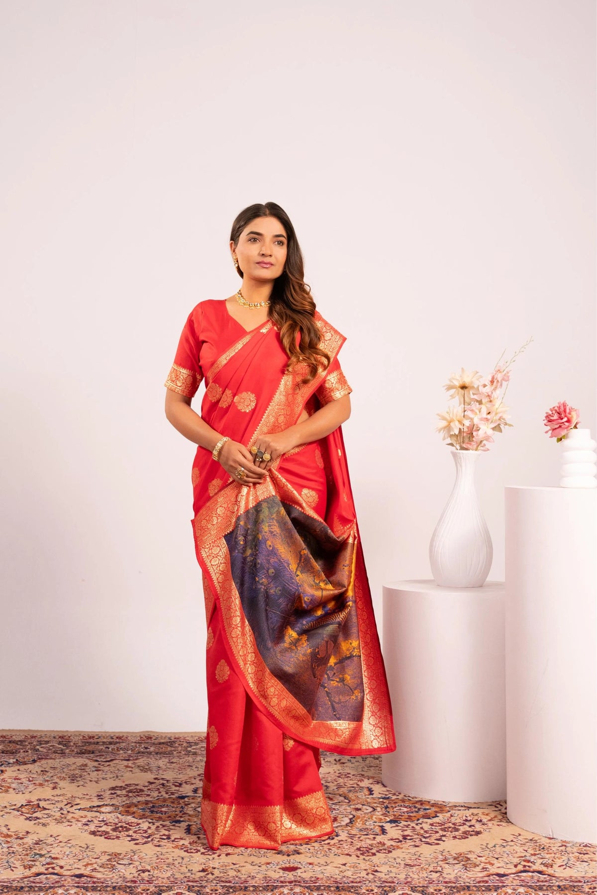 Woman in a red saree with gold border standing in a room with a white wall and decorative vase.