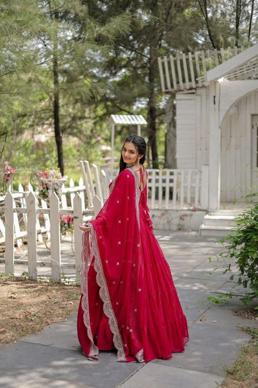 Woman in a red saree standing outdoors with greenery and a white structure in the background