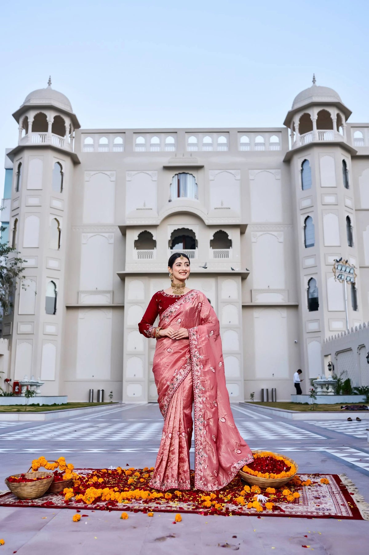 Woman in a pink saree standing in front of a building with decorative elements on the ground.