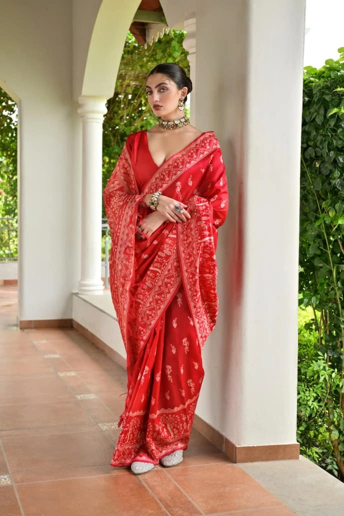 Woman in a red saree standing in an outdoor setting with greenery.